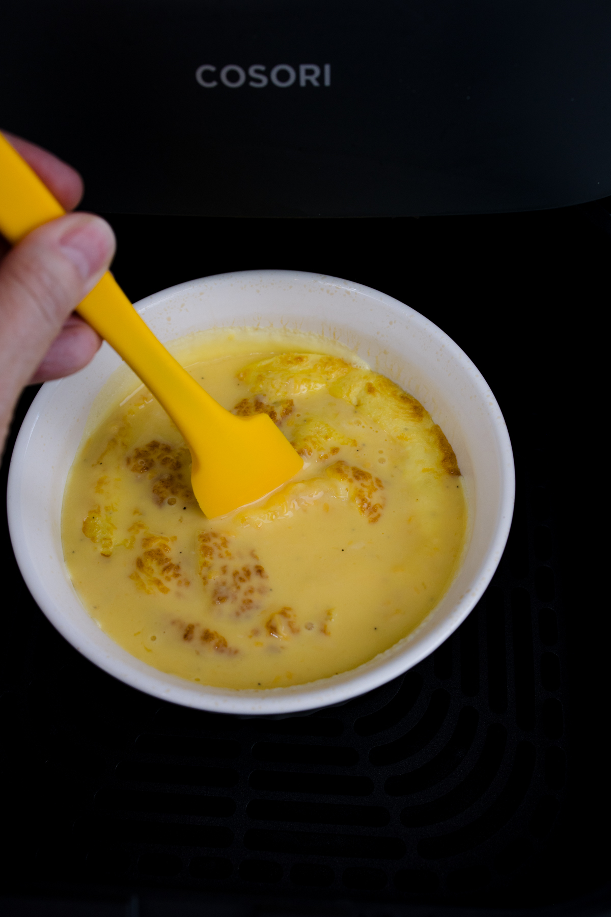 Close up view of half-cooked scrambled egg mixture in a white bowl in an air fryer, being stirred.