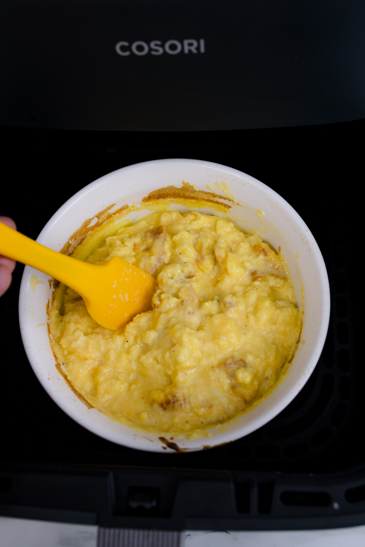 Close up view of cooked scrambled egg mixture in a white bowl in an air fryer, being stirred.