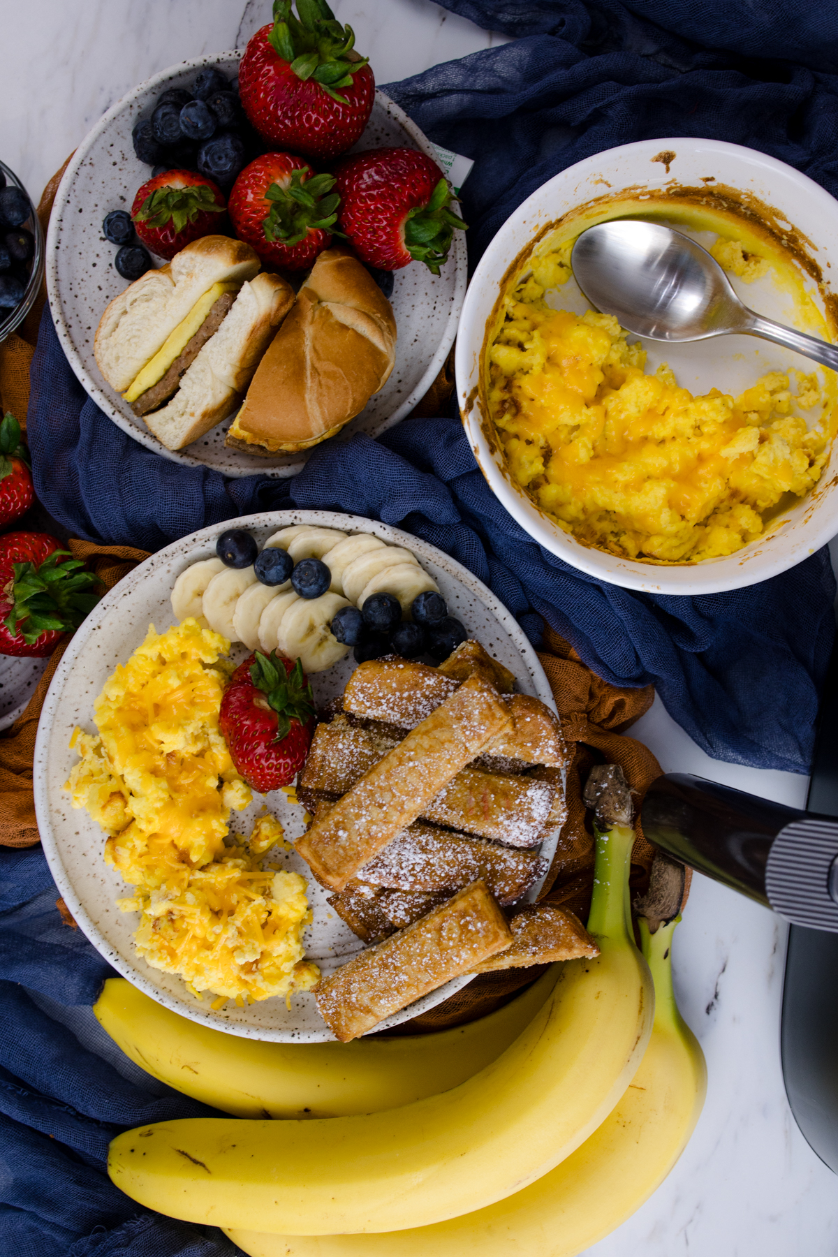 Air Fryer Scrambled Eggs on a white plate with other breakfast items, next to a bowl of scrambled eggs, and another plate with food on it.