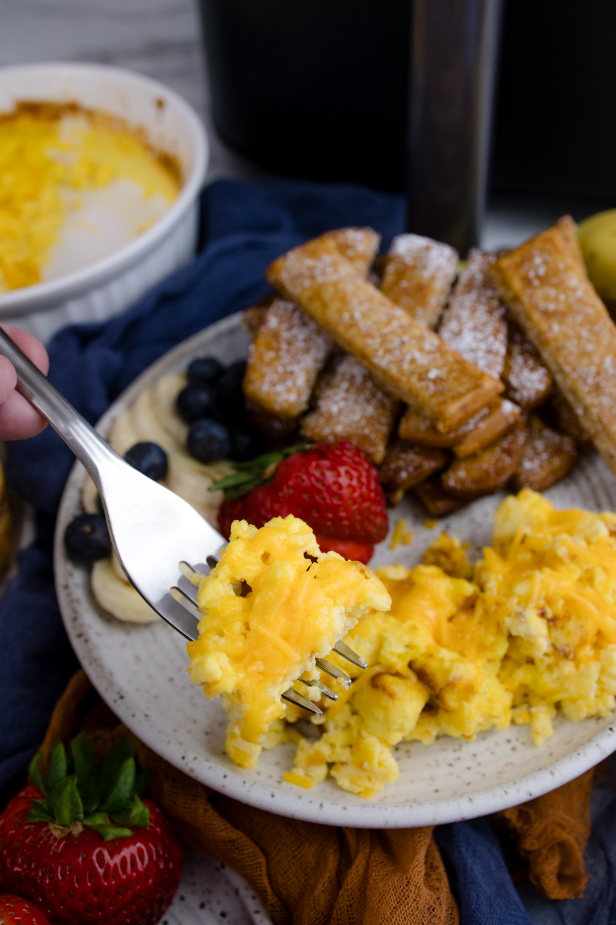 Air Fryer Scrambled Eggs on a fork being lifted up, over a white plate with more scrambled eggs and other breakfast items.