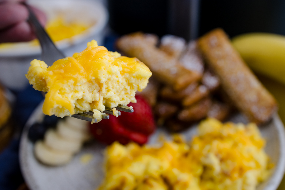 Air Fryer Scrambled Eggs on a fork being lifted up, over a white plate with more scrambled eggs and other breakfast items.