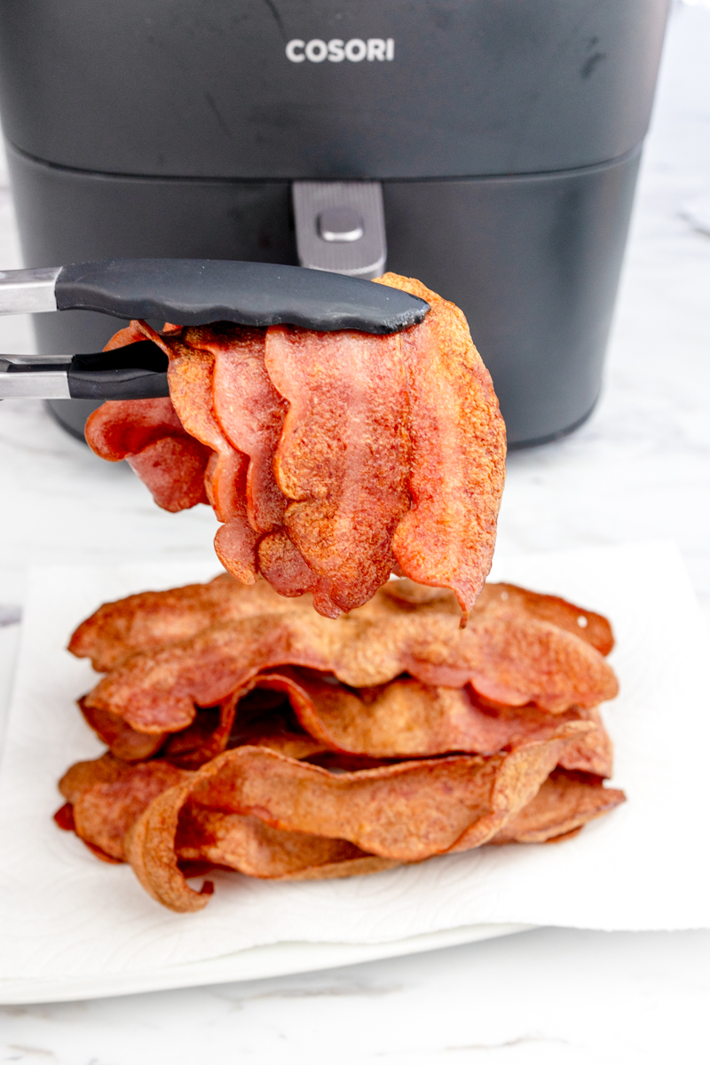 Close up shot of Air Fryer Turkey Bacon being lifted by a pair of tongs from a pile of bacon on a sheet of paper towel that's resting on a white plate. There is an air fryer in the background.