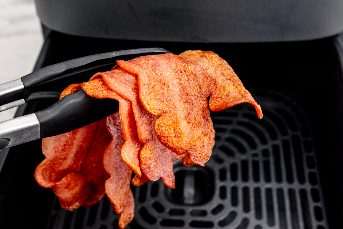 Close up shot of Air Fryer Turkey Bacon being lifted by a pair of tongs from an air fryer basket.