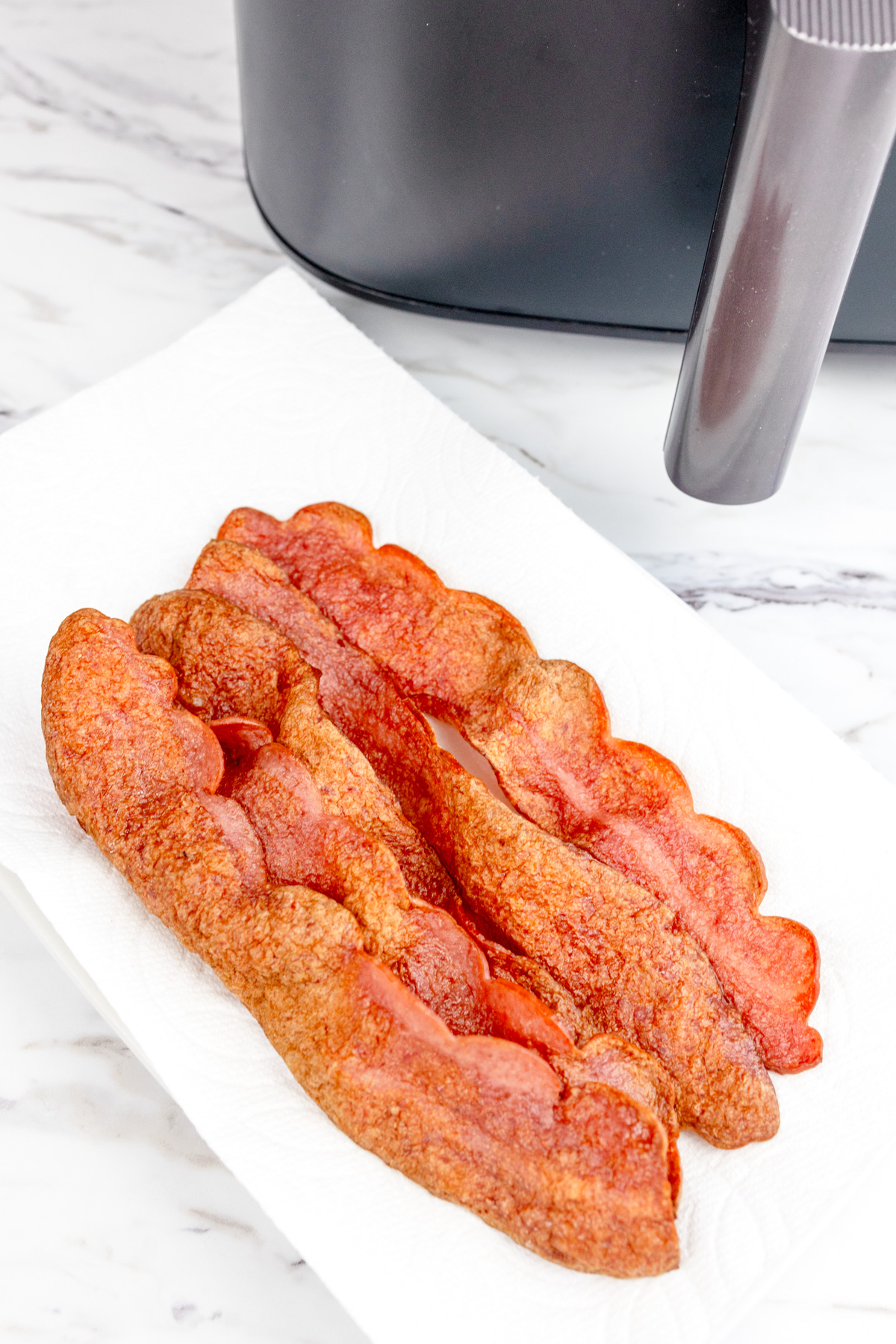 Close up shot of Air Fryer Turkey Bacon on a sheet of paper towel that's resting on a white plate. There is an air fryer in the background.