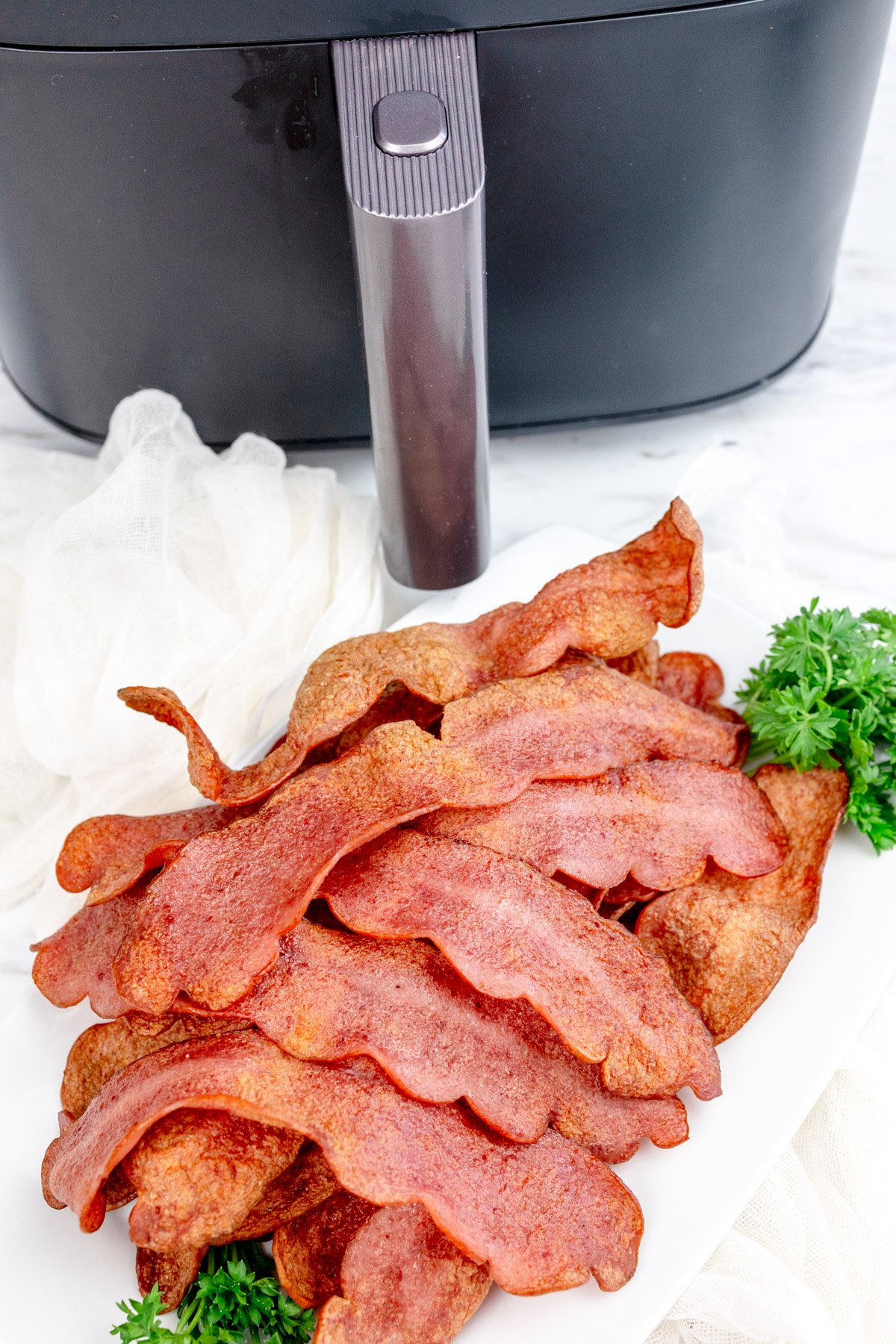 Close up shot of Air Fryer Turkey Bacon on a sheet of paper towel that's resting on a white plate with a leafy green garnish. There is an air fryer in the background.