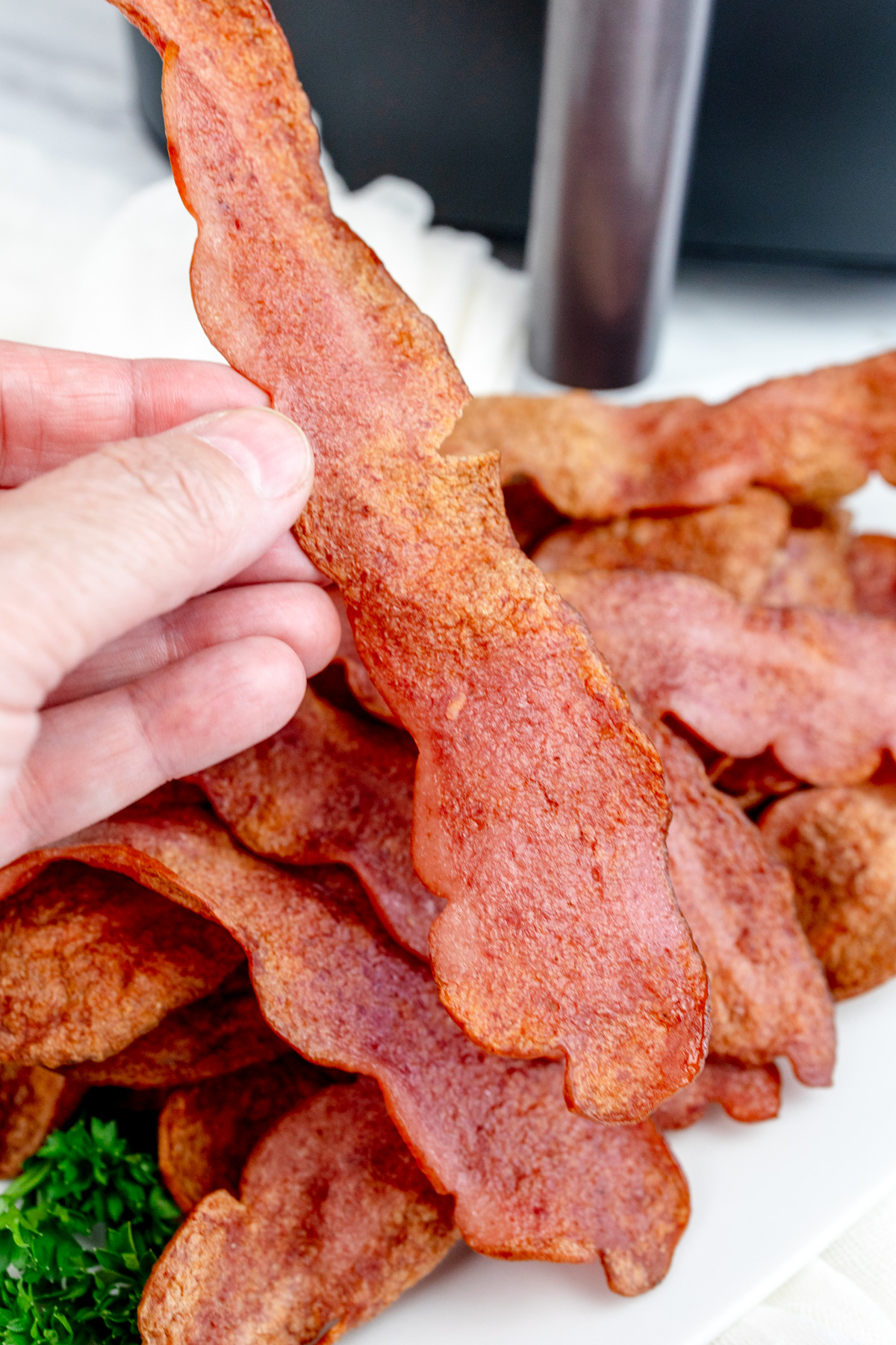 Close up shot of a hand lifting a slice of Air Fryer Turkey Bacon from a sheet of paper towel that's resting on a white plate. There is an air fryer in the background.