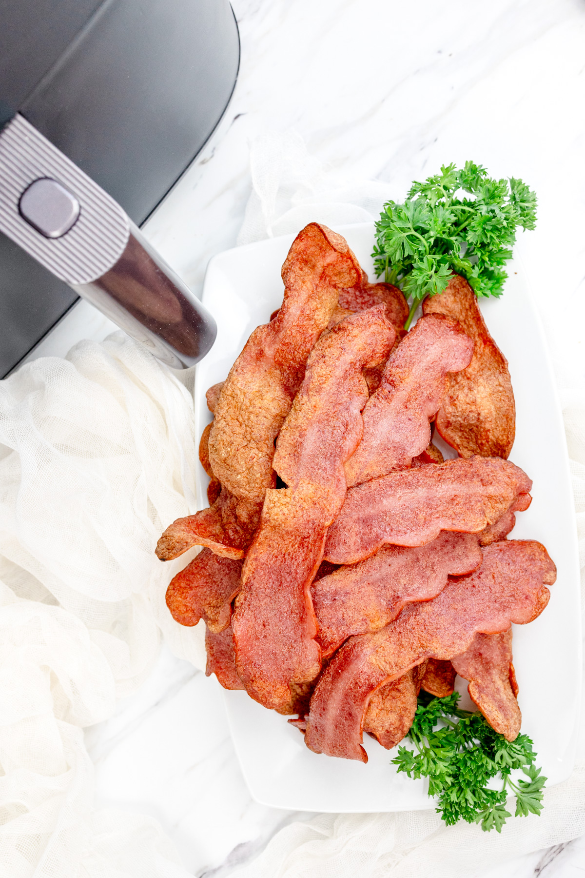 Close up shot of Air Fryer Turkey Bacon on a sheet of paper towel that's resting on a white plate with a leafy green garnish. There is an air fryer in the background.