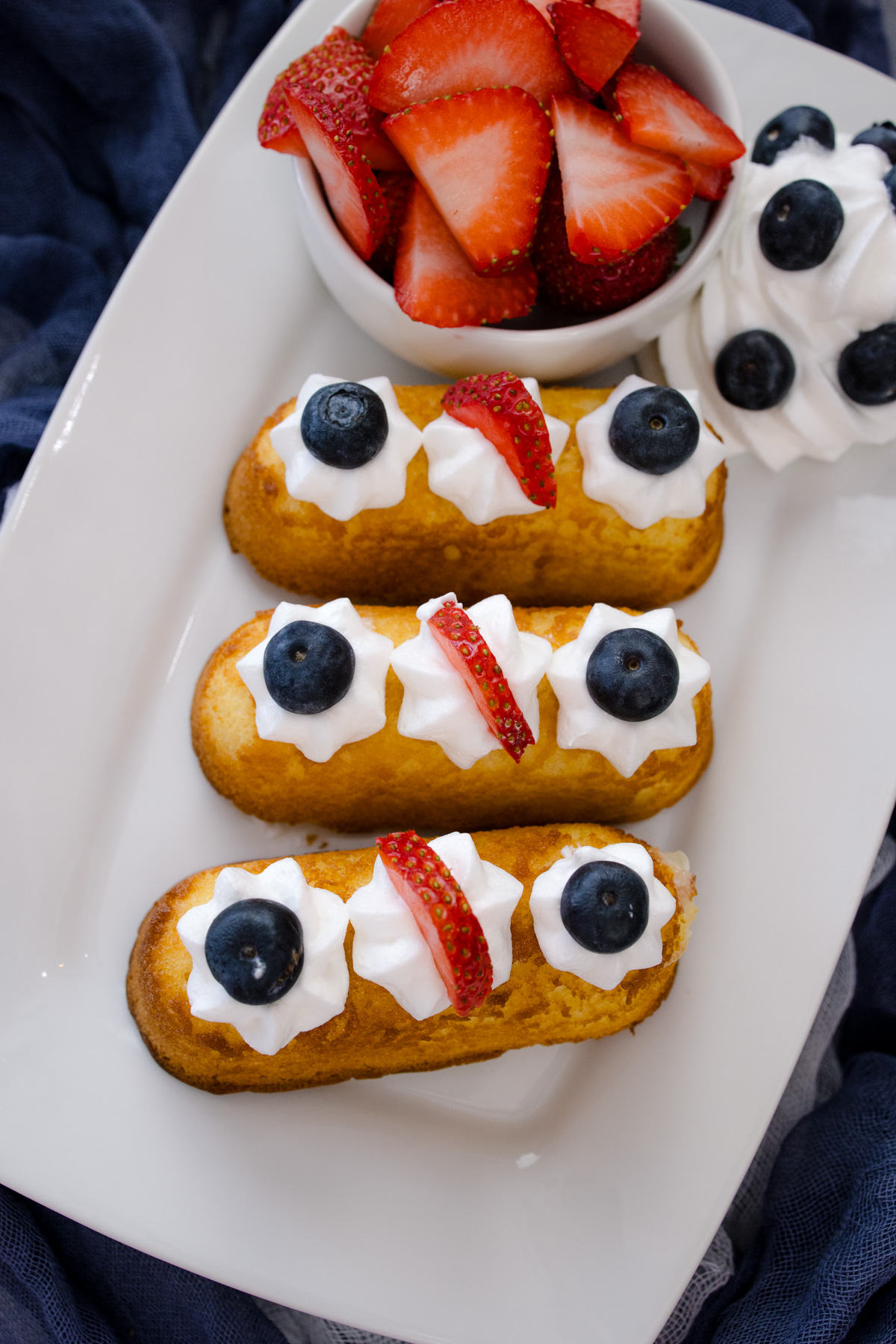 Air Fryer Twinkies topped with whipped cream and berries, on a white serving plate that has a bowl of berries on it.