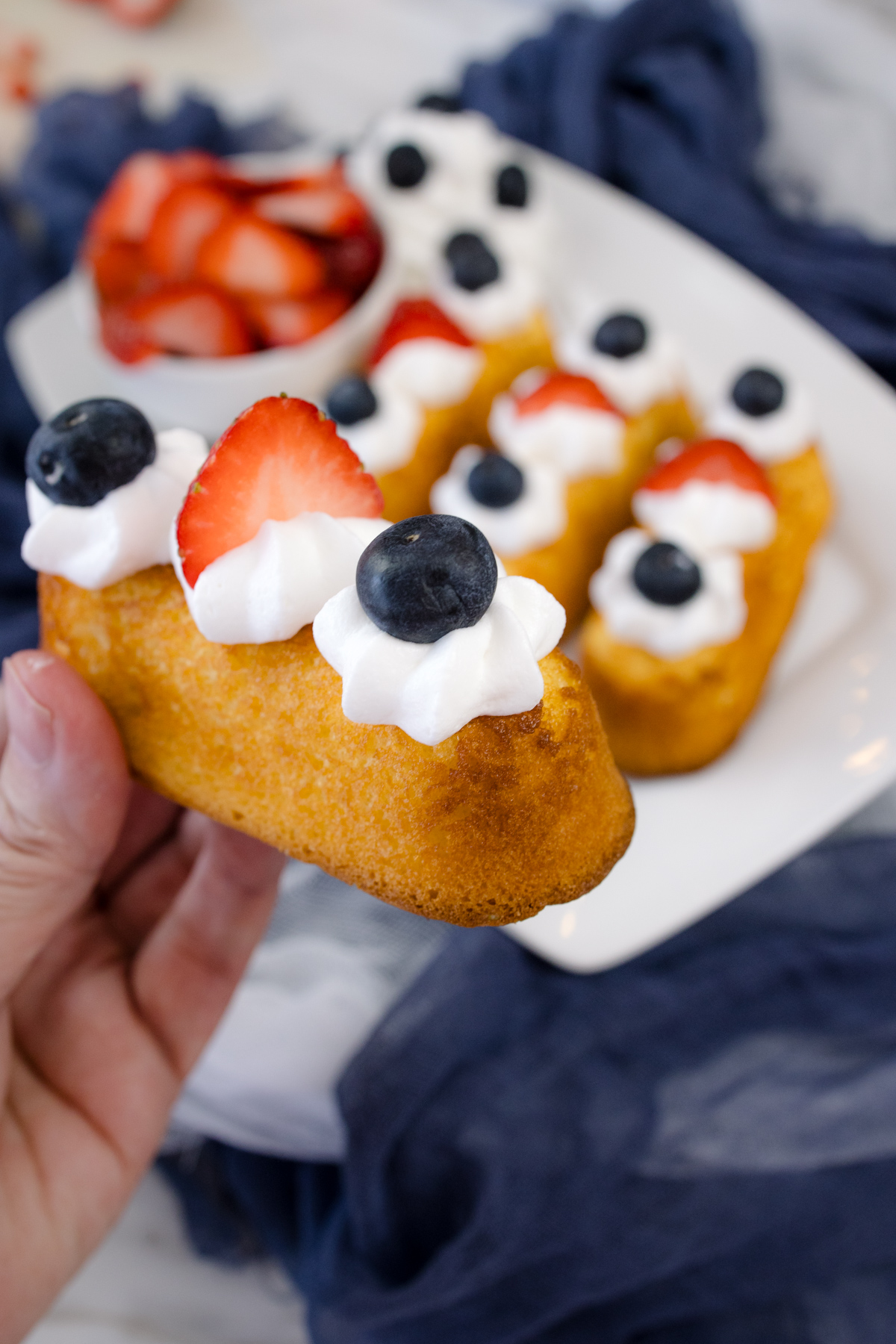 A hand holding up an Air Fryer Twinkie that's been topped with whipped cream and berries, over a white serving plate with other Twinkies on it.