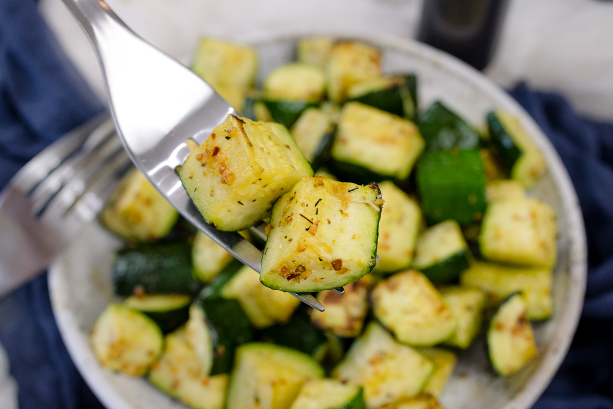 A fork carrying air fried zucchini over a plate with more zucchini in it.