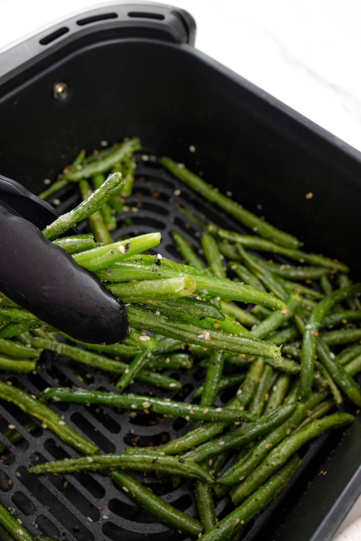 Tongs stirring green beans in an air fryer basket.