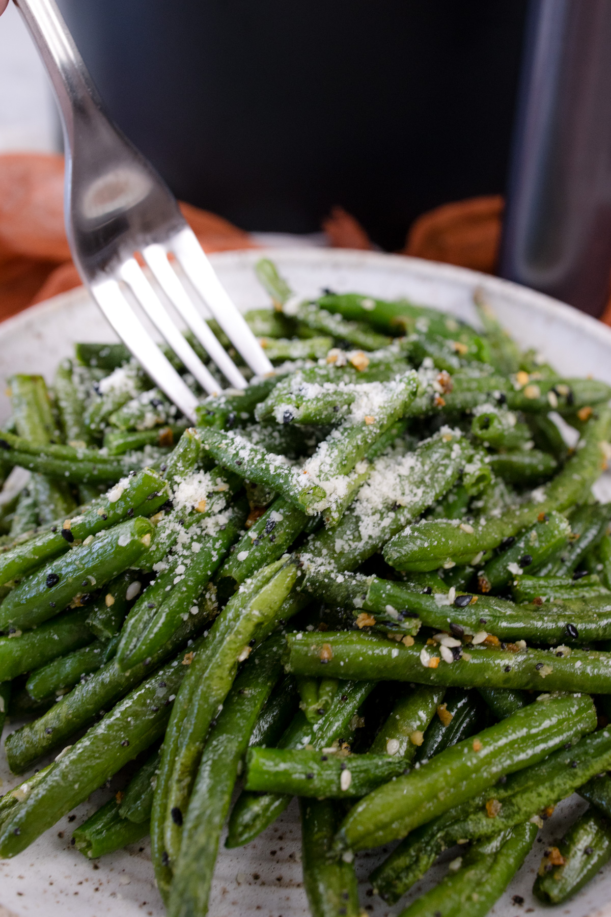 Air fryer frozen green beans on a white plate with a metal fork digging into it, and there's an air fryer in the background.