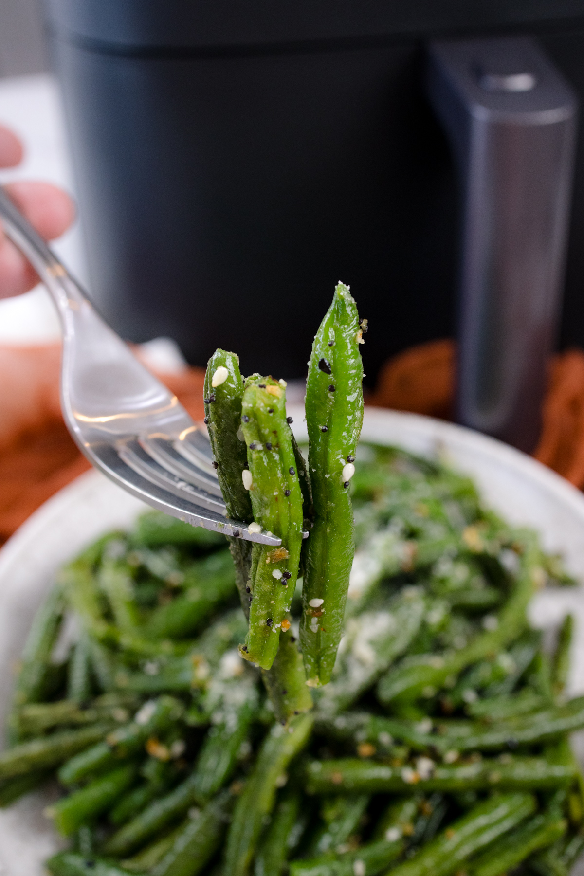 A fork lifting air fried green beans from a plate full in front of an air fryer.