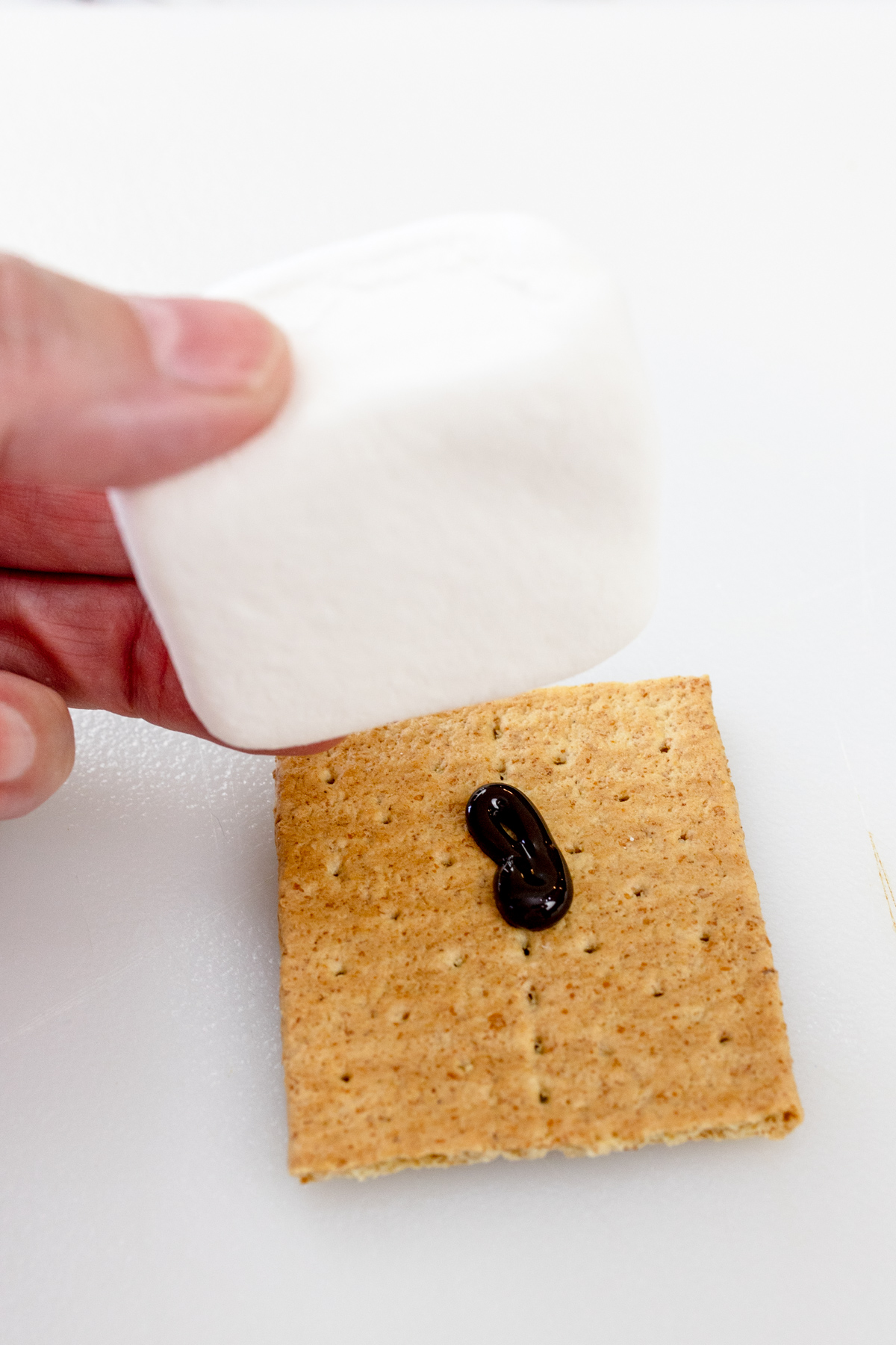 A hand placing a large white marshmallow on top of a graham cracker that has a bit of chocolate syrup on it.