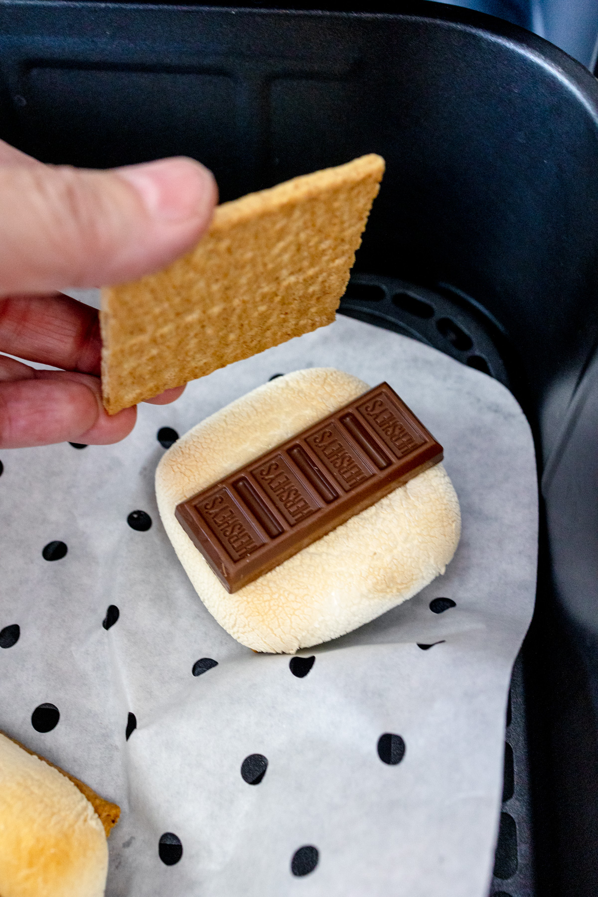 A hand placing a graham cracker on top of a piece of chocolate on a toasted marshmallow inside an air fryer basket lined with parchment paper.