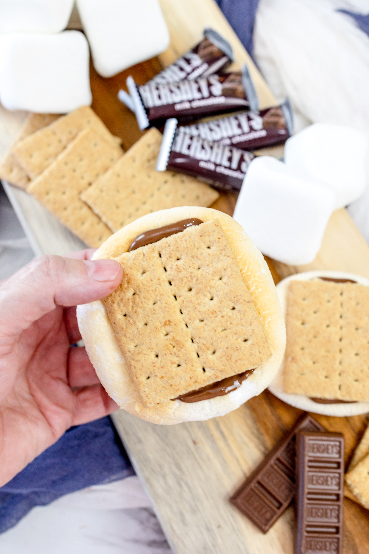 A hand holding an Air Fryer Smore over some Air Fryer Smores and the ingredients needed to make them on a wooden surface.