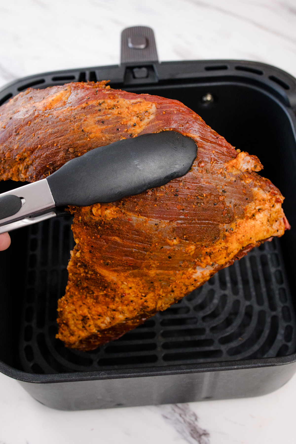 Seasoned Tri Tip Roast being held with tongs over an air fryer basket.