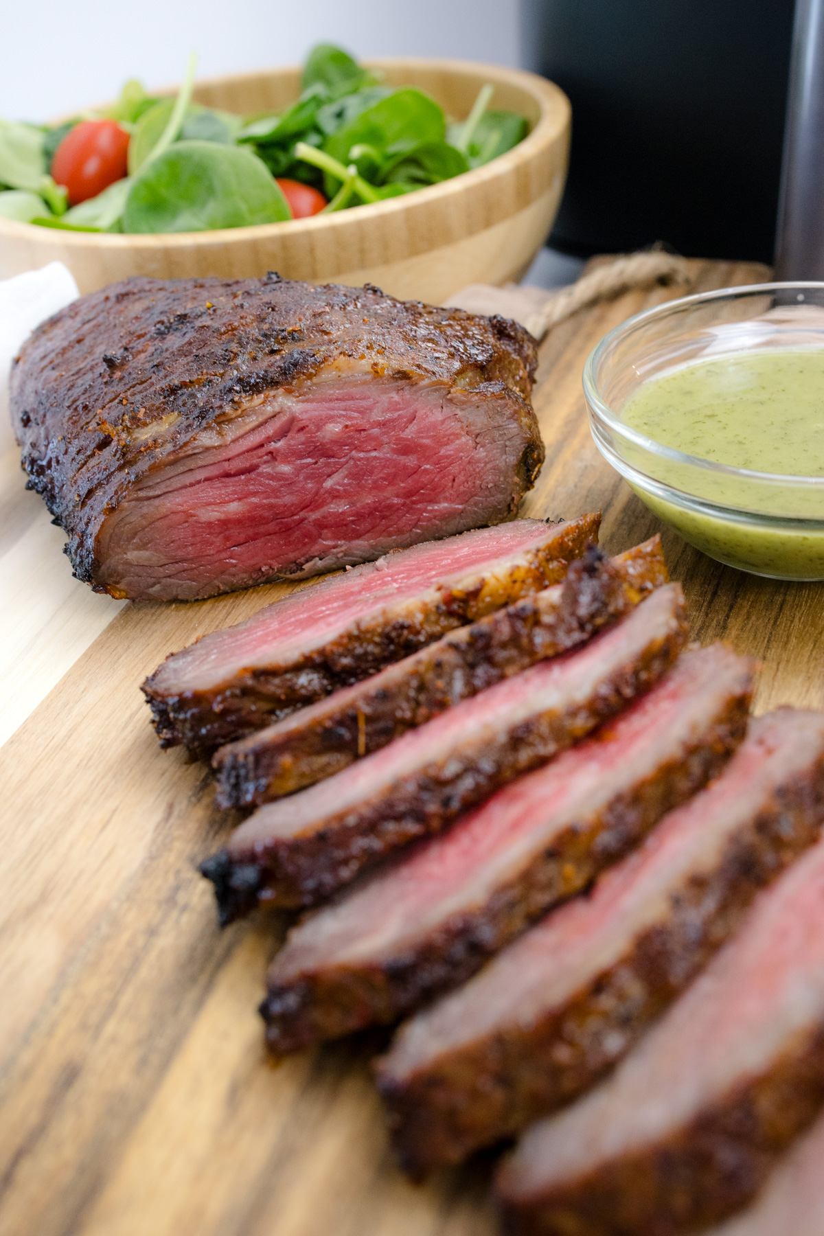 Medium rare Air Fried Tri Tip, in slices on a wooden surface, next to bowls of salad and sauce.