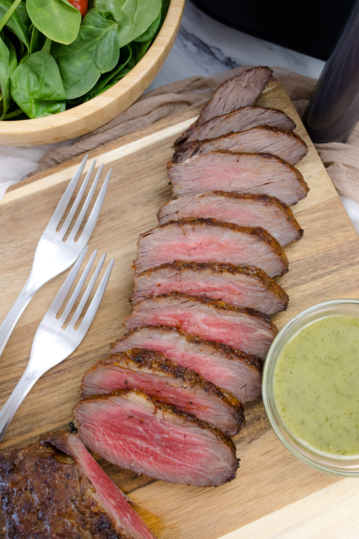 Medium rare Air Fried Tri Tip, in slices on a wooden surface, next to bowls of salad and sauce.