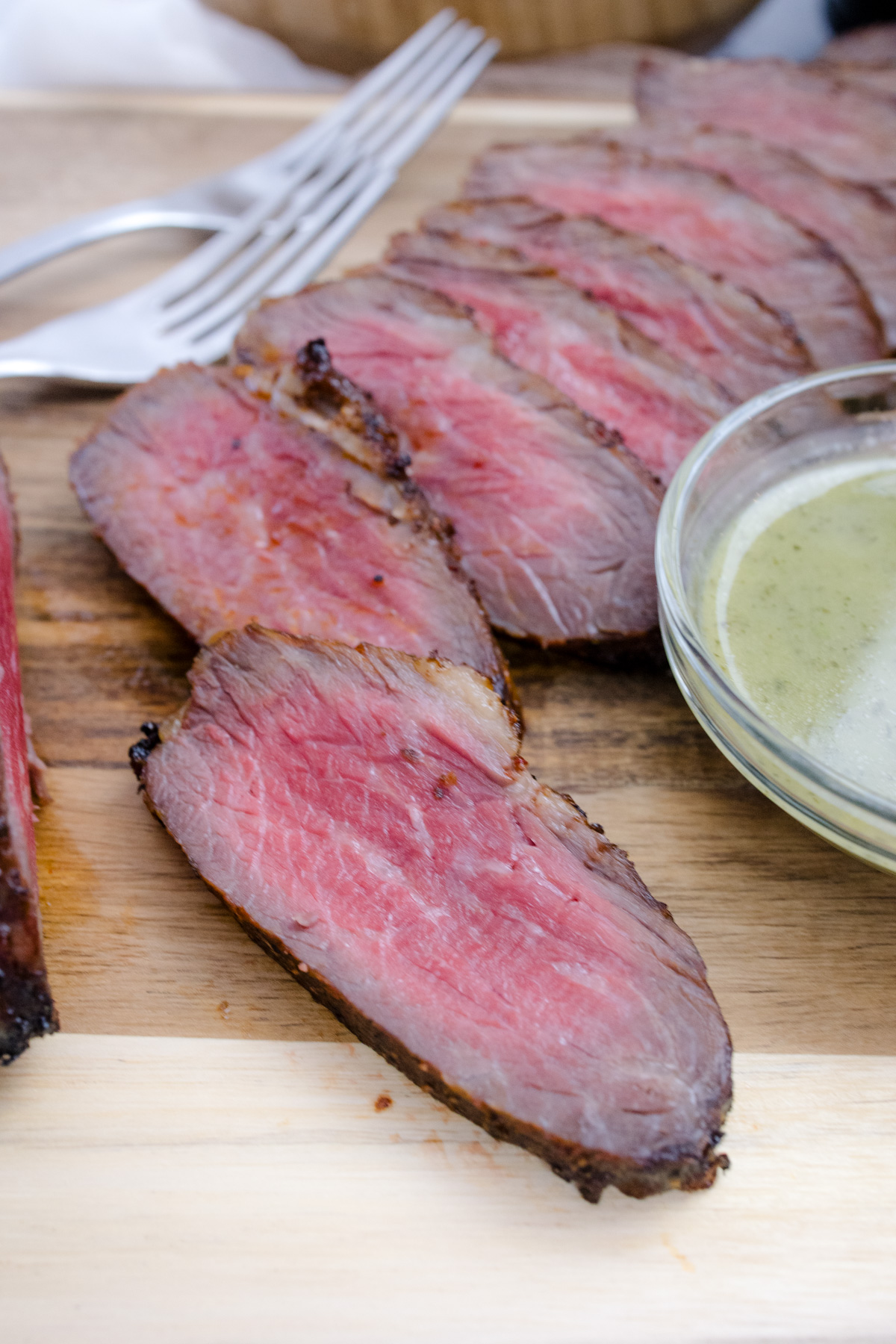 Medium rare Air Fried Tri Tip, in slices on a wooden surface, next to bowls of salad and sauce.