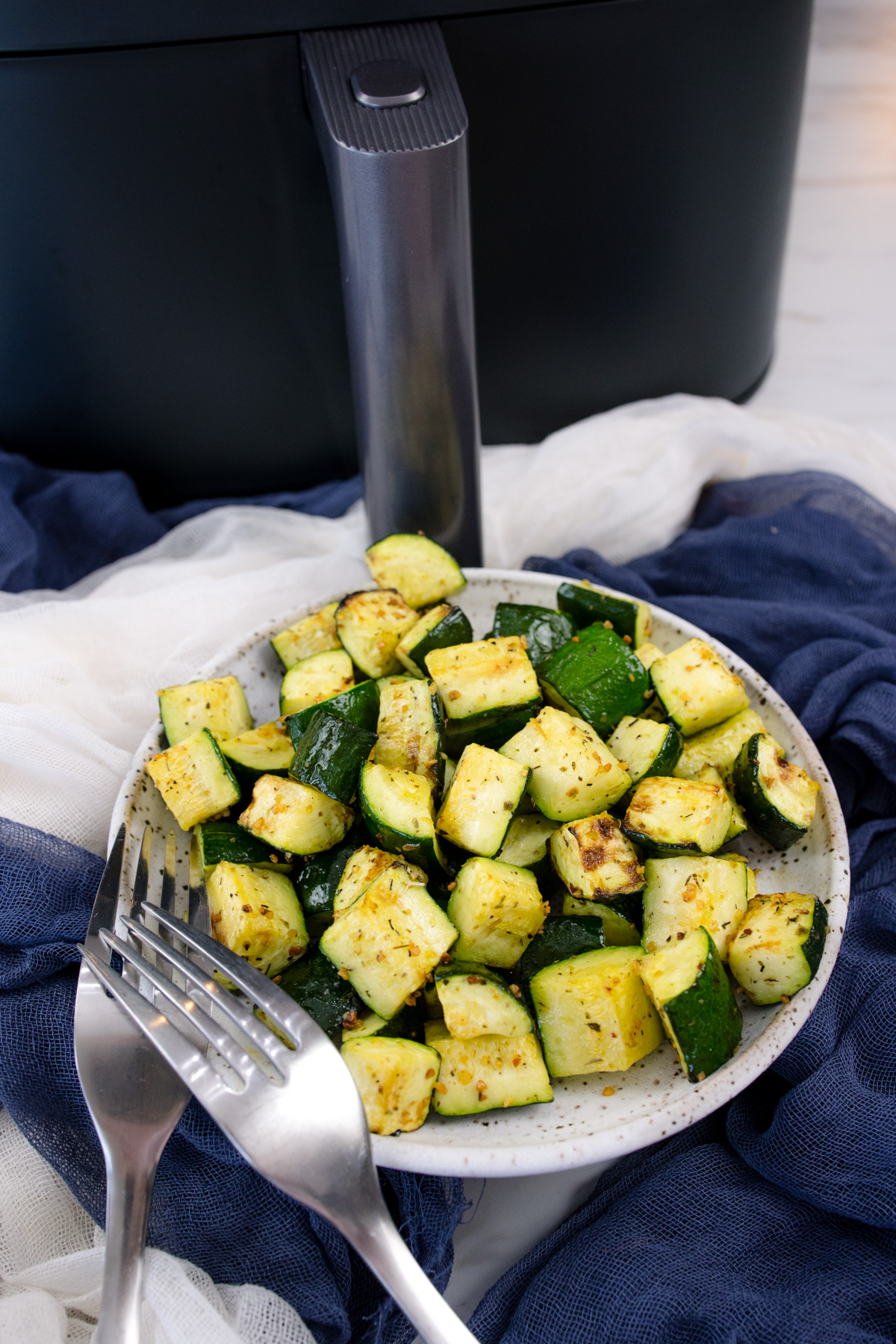 Air Fryer Zucchini on a white plate with a silver fork in the foreground and an air fryer in the background.