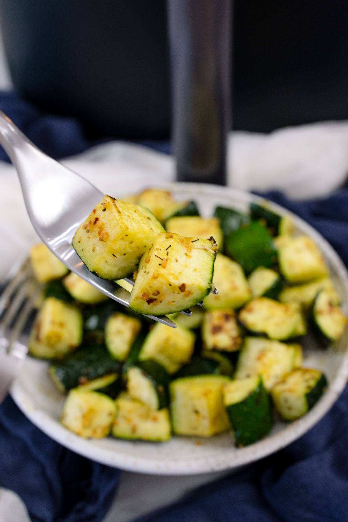 A fork carrying air fried zucchini over a plate with more zucchini in it.