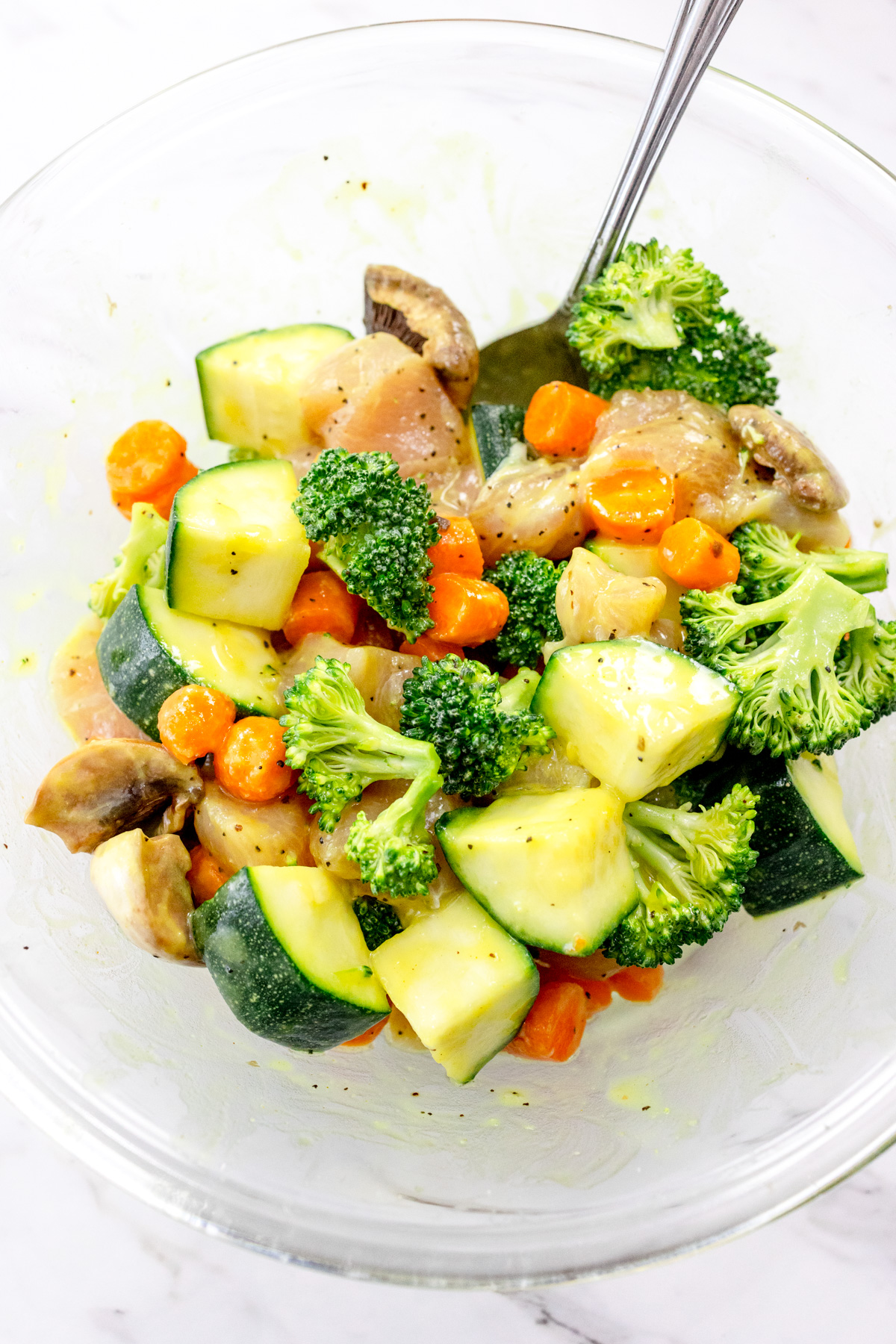 Chopped vegetables and chicken cubes being seasoned in a glass mixing bowl.