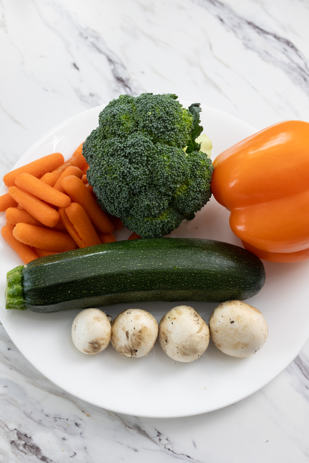 Raw vegetables (zucchini, carrots, orange bell pepper, broccoli, and 4 white mushrooms) on a white plate.