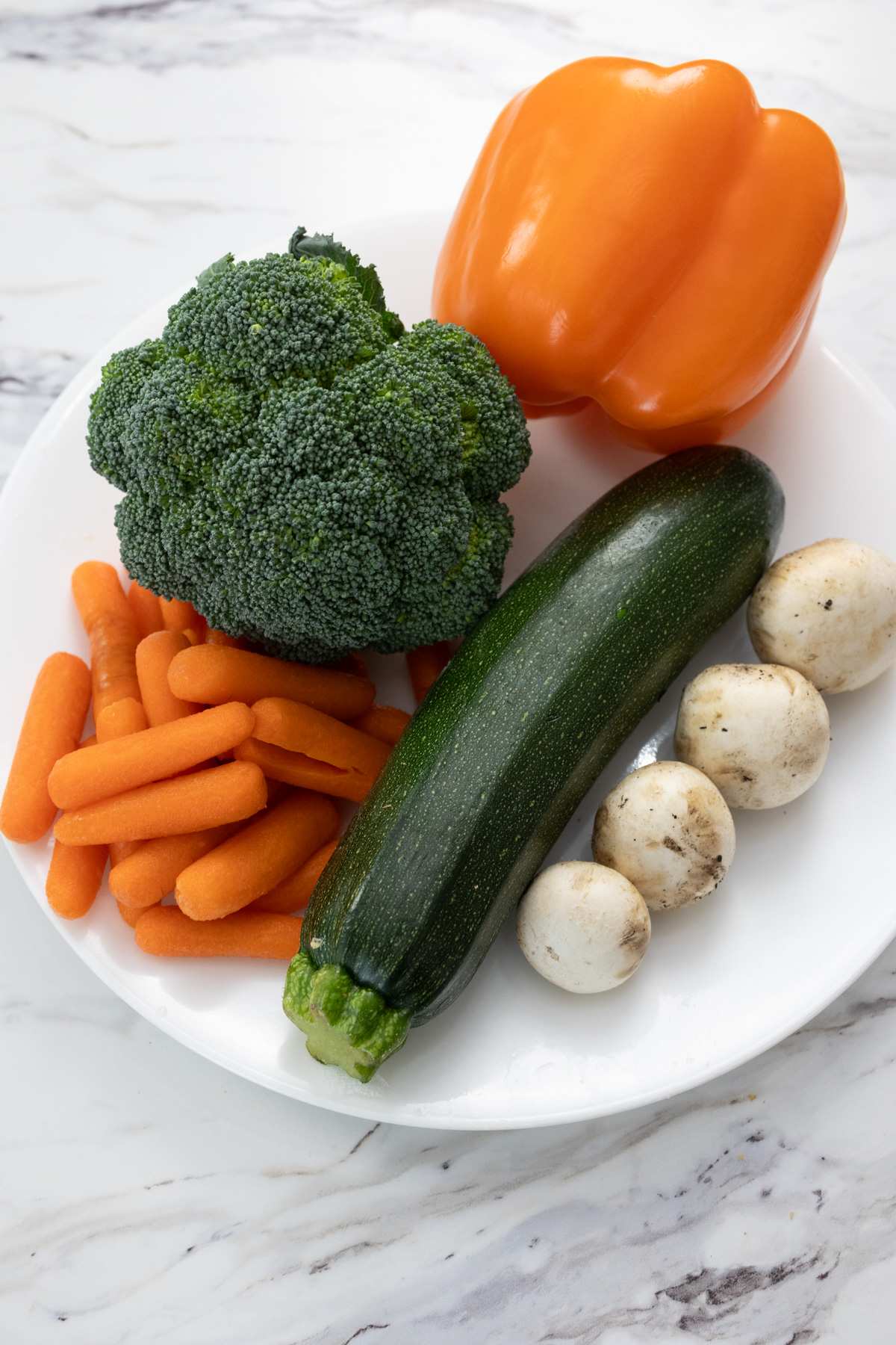 Raw vegetables (zucchini, carrots, orange bell pepper, broccoli, and 4 white mushrooms) on a white plate.