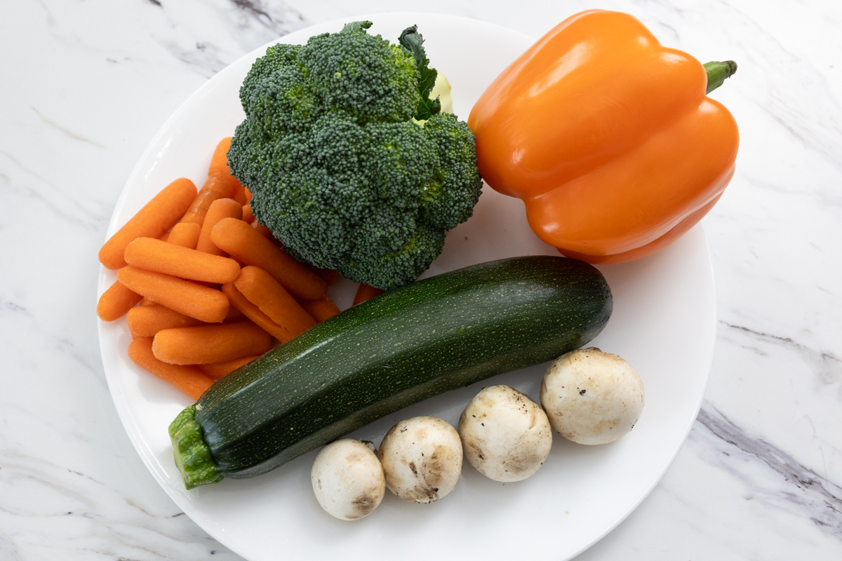 Raw vegetables (zucchini, carrots, orange bell pepper, broccoli, and 4 white mushrooms) on a white plate.