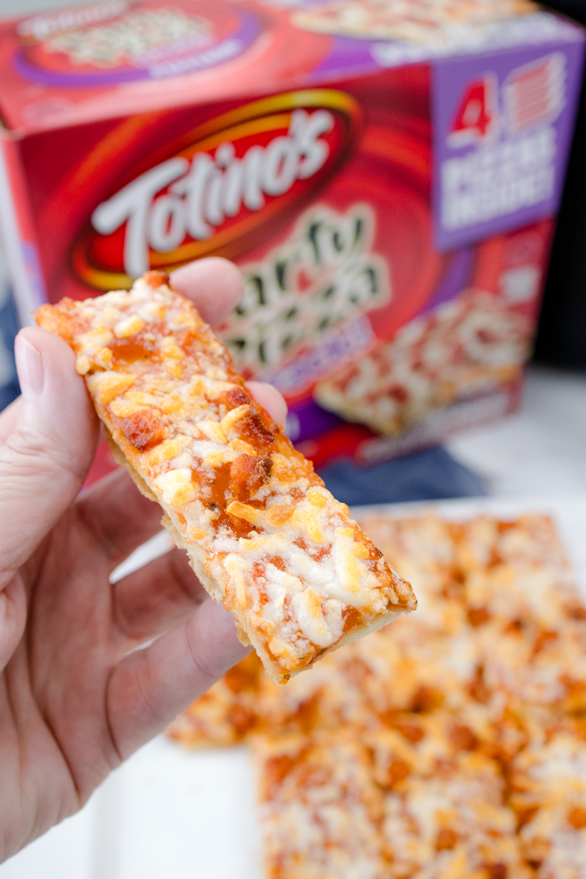 A hand holding a slice of pizza over a cooked air fryer pizza on a white surface in front of the packaging.