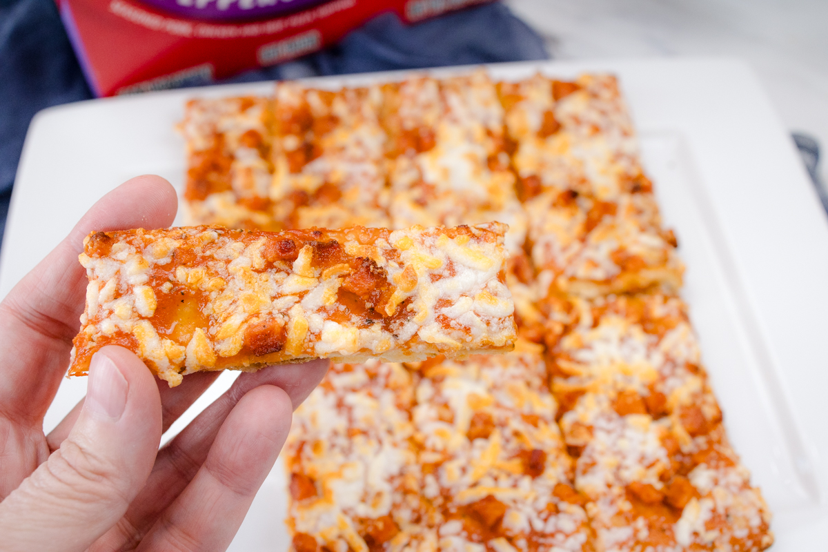 A hand holding a slice of pizza over a cooked air fryer pizza on a white surface in front of the packaging.