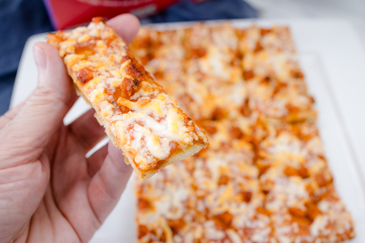 A hand holding a slice of pizza over a cooked air fryer pizza on a white surface in front of the packaging.