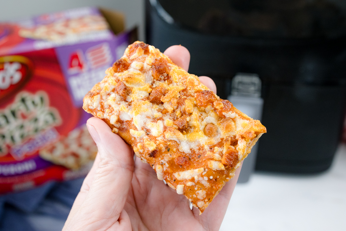 A hand holding a slice of pizza over, with the pizza box and air fryer in the background.